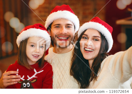 Young cheerful family of three in Santa hats with gifts taking selfie on phone in Christmas lights 119372654