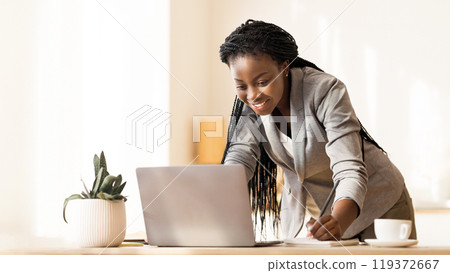 Young black female entrepreneur looking at laprop screen and taking notes while standing up near her desk, free space Young black female entrepreneur looking at laprop screen and taking notes while standing up near her desk, free space 119372667