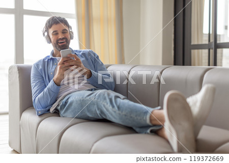 A man is lying on a grey couch with his legs extended and wearing headphones. He is smiling and looking at his phone. 119372669