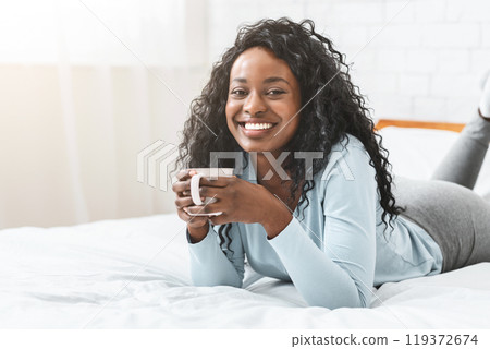 Young african woman enjoying aromatic coffee in bed, smiling at camera, free space 119372674