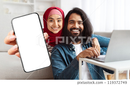 A cheerful Indian muslim couple sits closely together on a sofa, with one partner holding a phone and the other working on a laptop. They enjoy a relaxed moment at home while smiling at the camera. 119372749