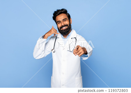 Indian doctor with a beard is smiling and making a call me gesture with his left hand while pointing his right index finger forward. He is standing in front of a blue background. 119372803