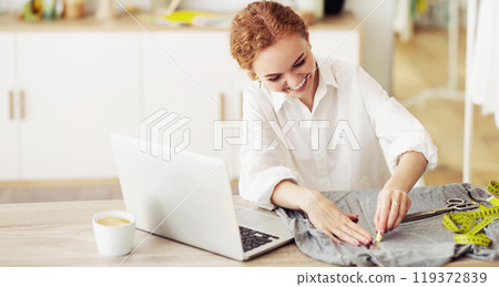 A woman smiles as she marks fabric with chalk, sitting at a table with a laptop and a cup of coffee in her home studio. A woman smiles as she marks fabric with chalk, sitting at a table with a laptop and a cup of coffee in her home studio. 119372839