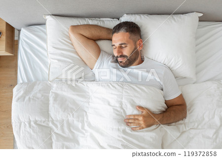 A bearded man is sleeping in a bed with white bedding. He is wearing a white t-shirt and has his arm behind his head. The bed has a white duvet and two white pillows, top view 119372858