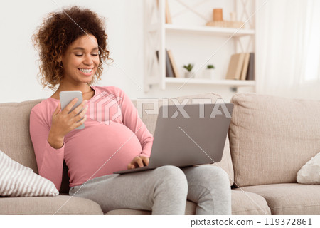 Happy expectant woman using technology devices, laptop and cellphone, resting on sofa, copy space Happy expectant woman using technology devices, laptop and cellphone, resting on sofa, copy space 119372861