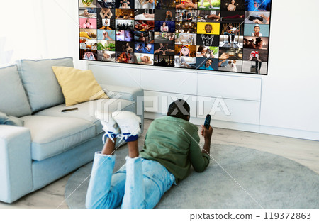 A man is lying on the floor of a living room, watching television. The television is displaying a grid of various images, and the man is holding a remote control in his hand. 119372863