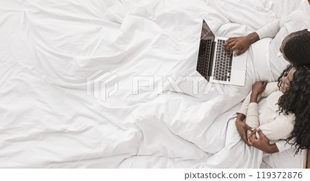 Afro couple using laptop in bed, cuddling and watching movies, top view 119372876