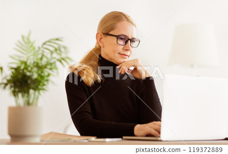 A woman wearing glasses is working on her laptop in a well-lit office. 119372889