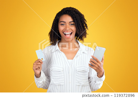 Online Shopping. Excited afro girl holding credit card and smartphone, looking at camera over yellow background 119372896