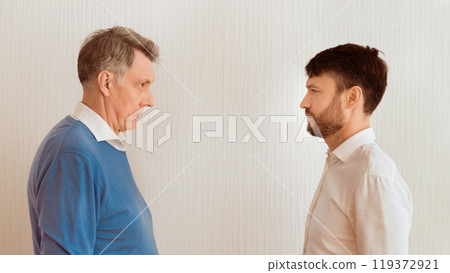 Conflict Between Generations. Elderly Man And Mature Son Looking Severely At Each Other Standing Face-To-Face Against White Wall Indoor. Panorama 119372921