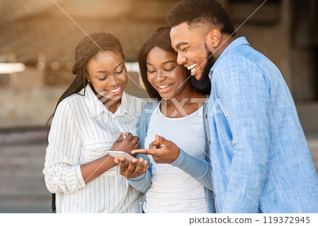 Funny content. Cheerful african american friends watching videos on smartphone, spending time together outdoors Funny content. Cheerful african american friends watching videos on smartphone, spending time together outdoors 119372945