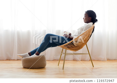 Relax at home. African american girl resting in wicker armchair and drinking coffee against window, free space Relax at home. African american girl resting in wicker armchair and drinking coffee against window, free space 119372947