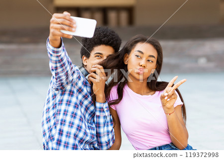 A teen black couple takes a selfie in front of a blurry background, likely outdoors in a city. Guy is holding the phone up, while the girl is making a funny face and giving the peace sign. 119372998