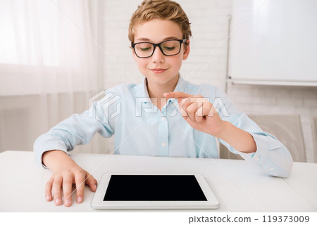 This image shows a young boy wearing glasses sitting at a white table. He is using a tablet and is pointing at the screen with his index finger. He appears to be focused on the device 119373009