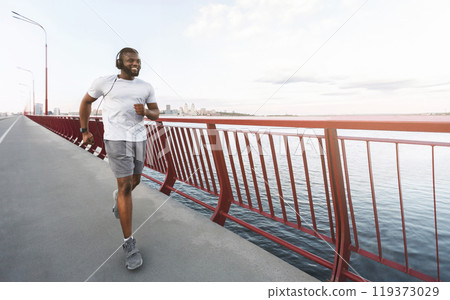 A man in headphones runs along a bridge overlooking a city skyline and a body of water. A man in headphones runs along a bridge overlooking a city skyline and a body of water. 119373029