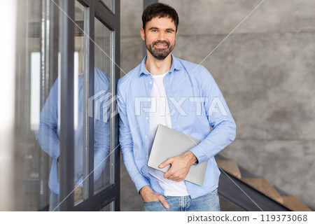 A man in a blue shirt and jeans stands in an office space, smiling and holding a laptop in his hands. He is looking directly at the camera, and his left hand is in his pocket. A man in a blue shirt and jeans stands in an office space, smiling and holding a laptop in his hands. He is looking directly at the camera, and his left hand is in his pocket. 119373068