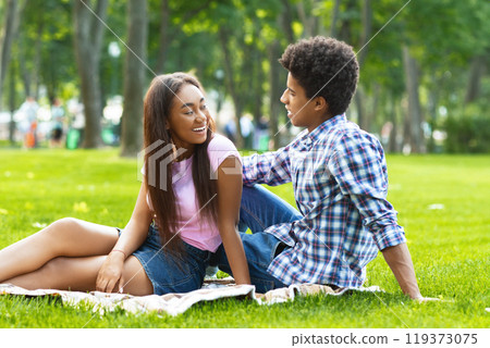 A teen black couple is enjoying a picnic date in a park. The girl is wearing a pink shirt and denim shorts and is sitting with her legs crossed, while guy is wearing a plaid shirt and jeans. 119373075