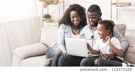 Planning vacation together. African american parents and their little daughter using laptop sitting on sofa at home. Planning vacation together. African american parents and their little daughter using laptop sitting on sofa at home. 119373108