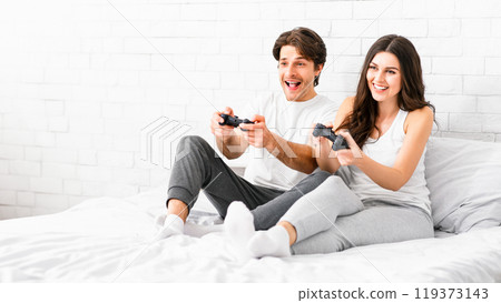 A couple sits on a white bed in a white room, playing video games with controllers in their hands. They both look excited and engaged in the game. 119373143