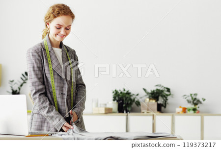 Tailor Concept. Smiling red-haired female seamstress standing at desk, cutting cloth, working with scissors tool 119373217