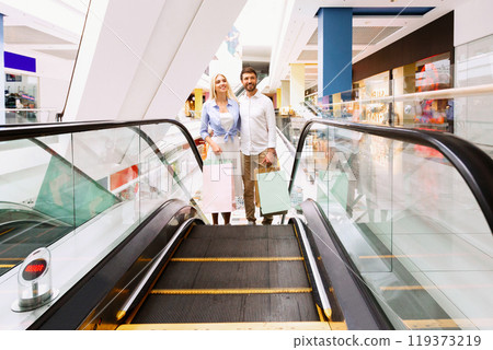A young couple, a man and a woman, stand on an escalator in a shopping mall, each holding a shopping bag. They are smiling and looking at the camera, having finished their shopping spree. 119373219