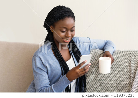 Leisure concept. Young african american woman texting on smartphone and drinking coffee, relaxing on comfortable sofa at home, free space Leisure concept. Young african american woman texting on smartphone and drinking coffee, relaxing on comfortable sofa at home, free space 119373225