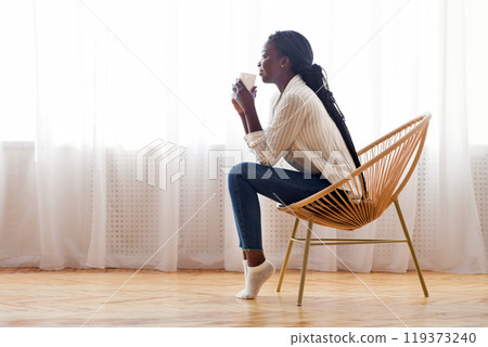 Time To Relax. African american girl sitting in wicker chair and drinking coffee against window at home, empty space Time To Relax. African american girl sitting in wicker chair and drinking coffee against window at home, empty space 119373240