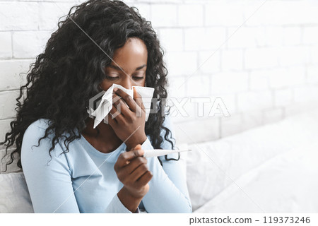 Sick african woman having fever, measuring body temperature with thermometer, blowing nose in bed, copy space Sick african woman having fever, measuring body temperature with thermometer, blowing nose in bed, copy space 119373246