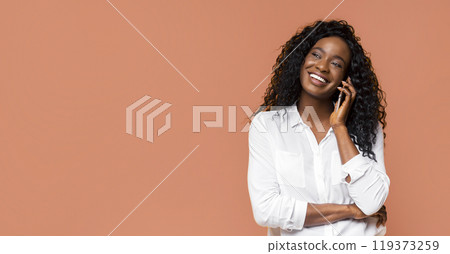 African American young woman with long curly hair is smiling while talking on a phone in front of a solid orange background. She is wearing a white shirt and has her arms crossed. African American young woman with long curly hair is smiling while talking on a phone in front of a solid orange background. She is wearing a white shirt and has her arms crossed. 119373259