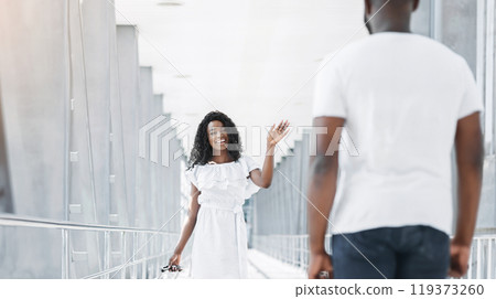Black woman in a white dress waves goodbye to a man in a white shirt in an airport terminal. The man is turned away from the camera, and the woman is looking directly at the camera with a smile. Black woman in a white dress waves goodbye to a man in a white shirt in an airport terminal. The man is turned away from the camera, and the woman is looking directly at the camera with a smile. 119373260