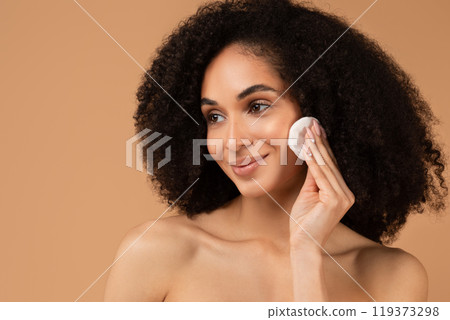 A young African American woman with curly hair smiles while applying makeup to her cheek with a cotton pad in her right hand. The background is a warm, light brown color. A young African American woman with curly hair smiles while applying makeup to her cheek with a cotton pad in her right hand. The background is a warm, light brown color. 119373298