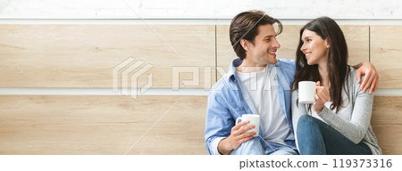 A couple is sitting on the floor in a kitchen, drinking coffee and talking. They both have relaxed expressions and are looking at each other lovingly, copy space 119373316