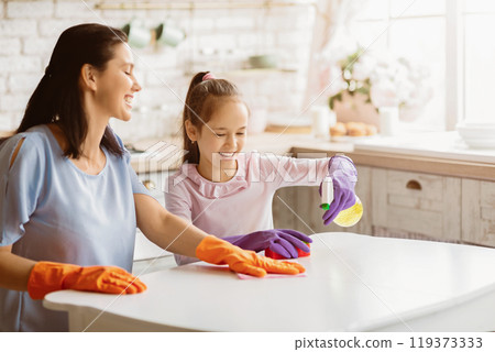 A mother and daughter are cleaning their kitchen table together. The mother is wearing orange gloves and wiping the table with a cloth, while the daughter is wearing purple gloves and spraying 119373333