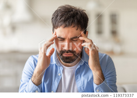 A man sits indoors with his eyes closed and hands pressed against his temples. He appears to be experiencing a headache or migraine. 119373337