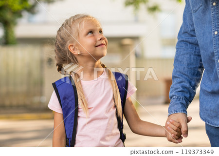 A young girl with blonde pigtails and a blue backpack walks to school holding the hand of her parent. She is looking up and smiling as they walk down the sidewalk. 119373349