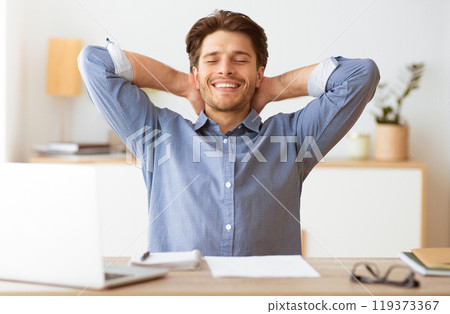 A relaxed man sits at his desk, leaning back with his arms behind his head, smiling and taking a break from work. He appears content and at ease. 119373367