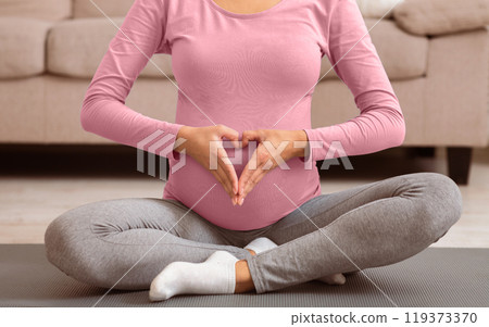 Pregnant Woman Holding Hands In Heart Shape On Her Belly, Sitting On The Floor At Home. Cropped view Pregnant Woman Holding Hands In Heart Shape On Her Belly, Sitting On The Floor At Home. Cropped view 119373370