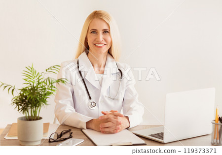A smiling female doctor sits at a desk in her office, ready to see her patients. She is wearing a white coat and stethoscope. 119373371