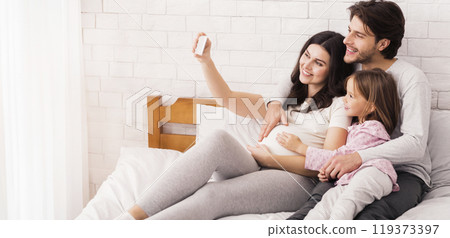 Pregnant Woman Taking Selfie With Her Husband And Daughter In Bed, Enjoying Spending Time At Home, Bonding In Bedroom 119373397