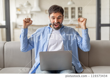A man sits on a couch, smiling widely and raising both fists in the air, while looking at a laptop screen. He appears to be celebrating a victory or success. A man sits on a couch, smiling widely and raising both fists in the air, while looking at a laptop screen. He appears to be celebrating a victory or success. 119373434