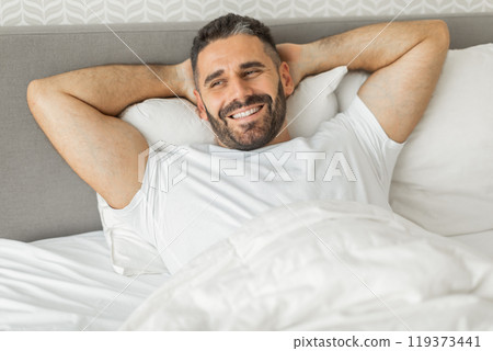 A man with a beard and short hair is lying in bed, smiling and looking directly at the camera. He is wearing a white t-shirt and his hands are behind his head, resting on a white pillow. A man with a beard and short hair is lying in bed, smiling and looking directly at the camera. He is wearing a white t-shirt and his hands are behind his head, resting on a white pillow. 119373441