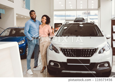 Car Buyers. Joyful Black Couple Hugging Showing Their New Auto Key Standing Near Automobile In Dealership Center. 119373456