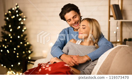 Cozy Christmas eve. Loving couple embracing near Christmas tree, resting on sofa Cozy Christmas eve. Loving couple embracing near Christmas tree, resting on sofa 119373470
