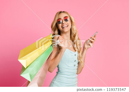 A happy woman with long blonde hair is wearing sunglasses, holding shopping bags and a smartphone. She is smiling broadly at the camera and appears to be enjoying her shopping experience. 119373547