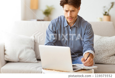 A man sits on a couch, working on a laptop and taking notes in a notebook. He appears focused and engaged in his work. A man sits on a couch, working on a laptop and taking notes in a notebook. He appears focused and engaged in his work. 119373552