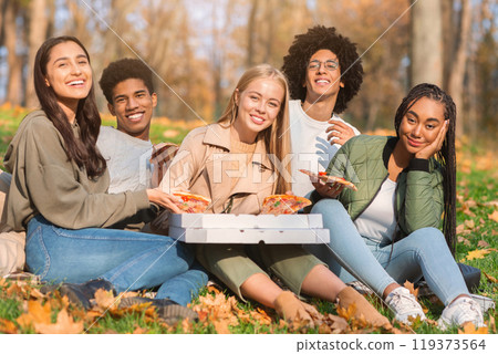 Group of positive young international friends with take away pizza sitting on ground at public park 119373564