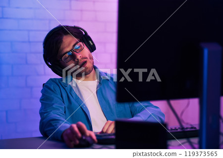 A young man wearing glasses and headphones is asleep at a computer desk. He is sitting in a chair with his head resting on his hand. The desk is lit by a pink and blue light A young man wearing glasses and headphones is asleep at a computer desk. He is sitting in a chair with his head resting on his hand. The desk is lit by a pink and blue light 119373565