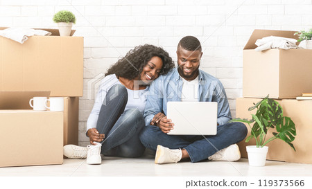 Happy african couple purchasing furniture for new apartment, sitting on floor and using laptop, panorama with free space Happy african couple purchasing furniture for new apartment, sitting on floor and using laptop, panorama with free space 119373566