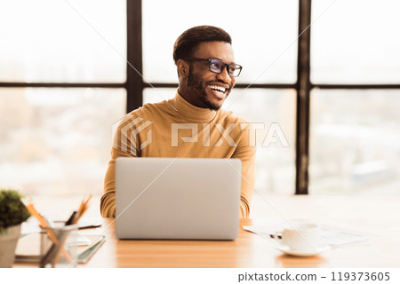Success At Work. Happy afro businessman sitting at desk by large window in modern office, looking aside at copyspace 119373605