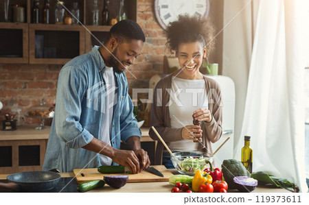 Laughing black couple preparing healthy salad together in loft kitchen. Young family cooking dinner 119373611
